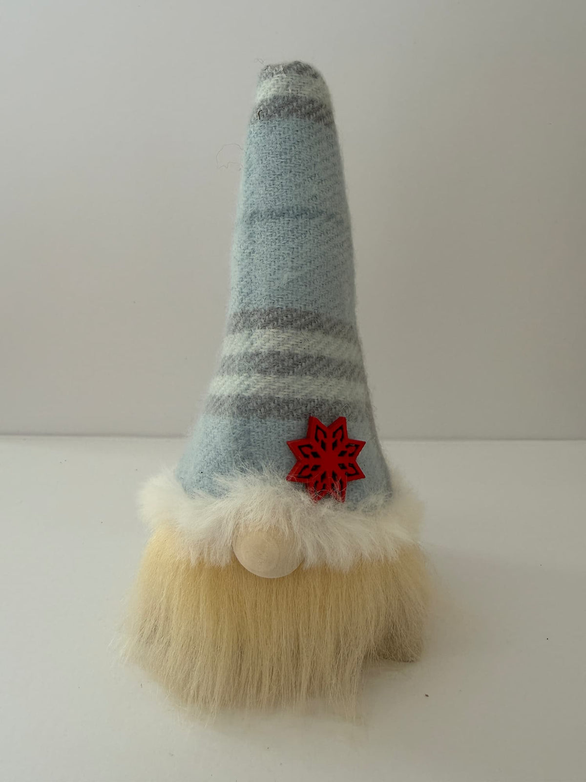 A small decorative Nordic gnome figurine with a tall pointed hat in light blue and gray stripes, a red snowflake ornament, a wooden nose and a fluffy cream beard on a plain light background. Handmade by Northern Nomes.