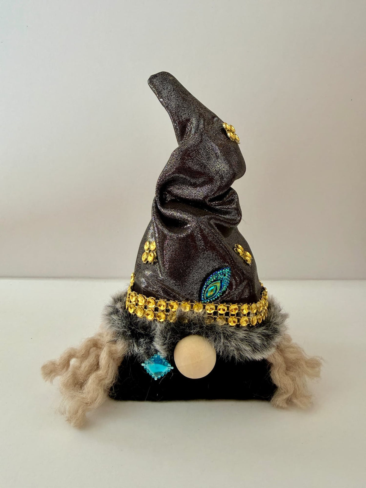A small witch hat gnome decoration with a dark black glittery hat, gold rhinestone trim, a faux fur band, a round wooden nose, and curly beige hair, photographed against a plain light background. Handmade by Northern Nomes.