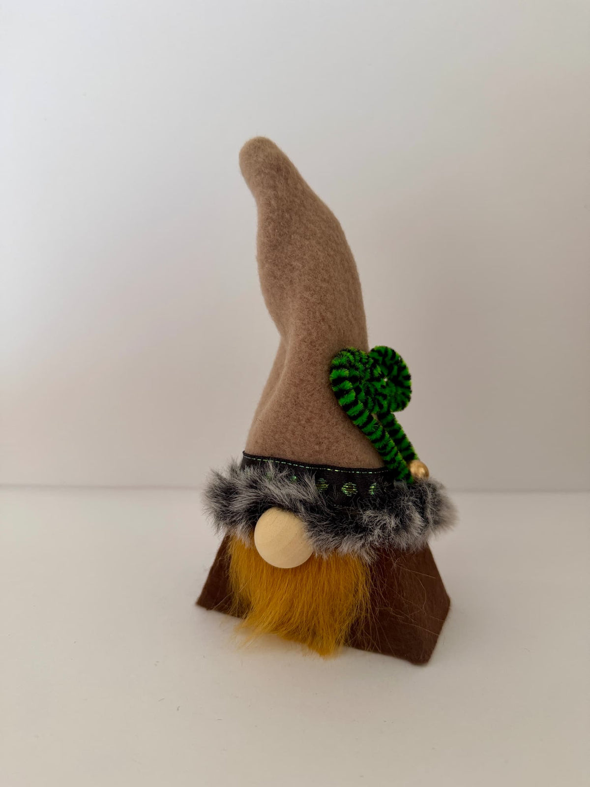 A small handmade gnome decoration with a tall tan hat, ginger beard, brown body, gray faux-fur trim, and a green decorative accent, photographed on a plain light background. Made by Northern Nomes.