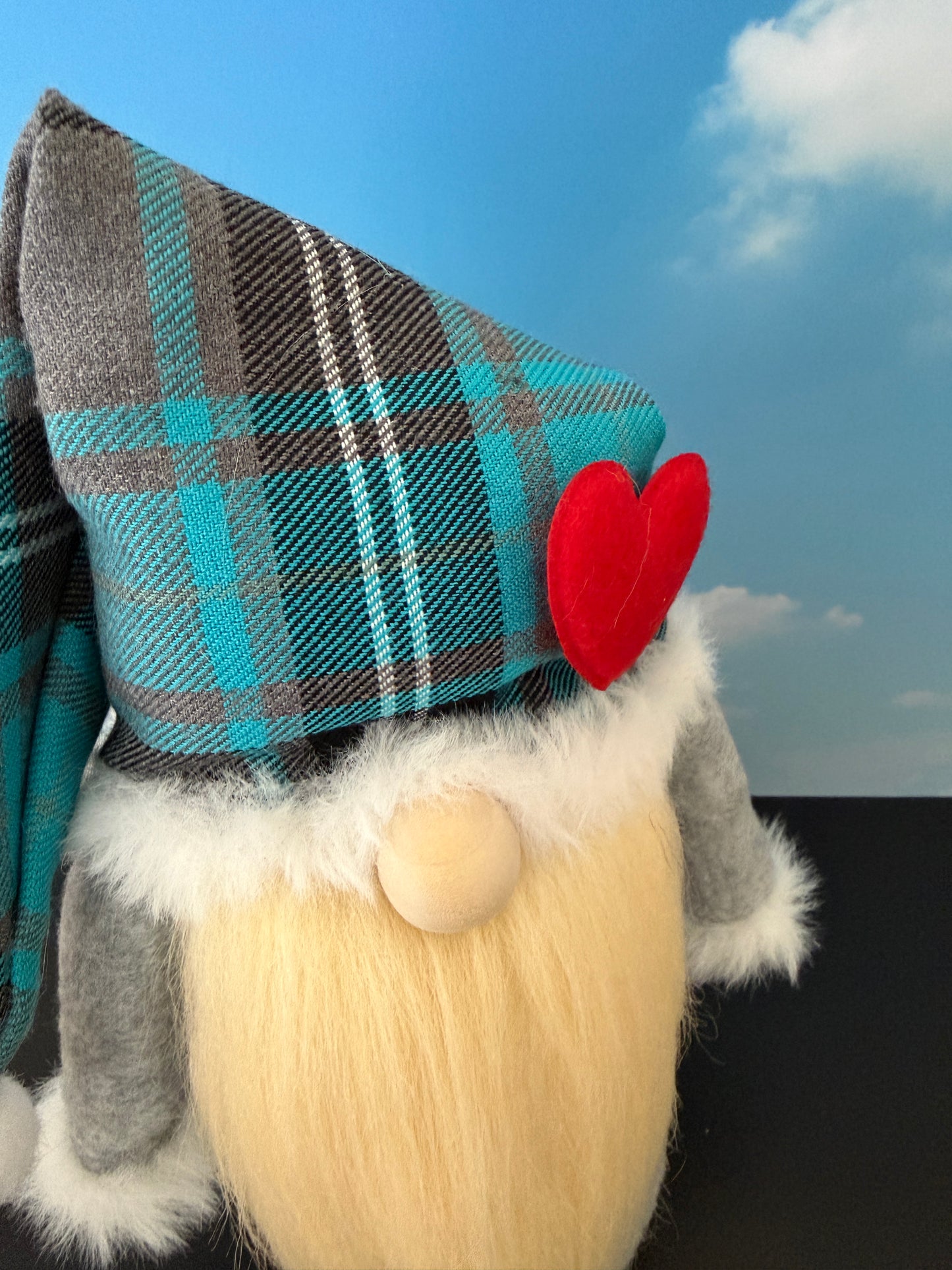 A close-up of a Northern Nomes handmade gnome wearing a teal, gray, and white plaid pointed nightcap with a red heart accent, a cream beard, and gray sleeves, set against a blue sky backdrop.