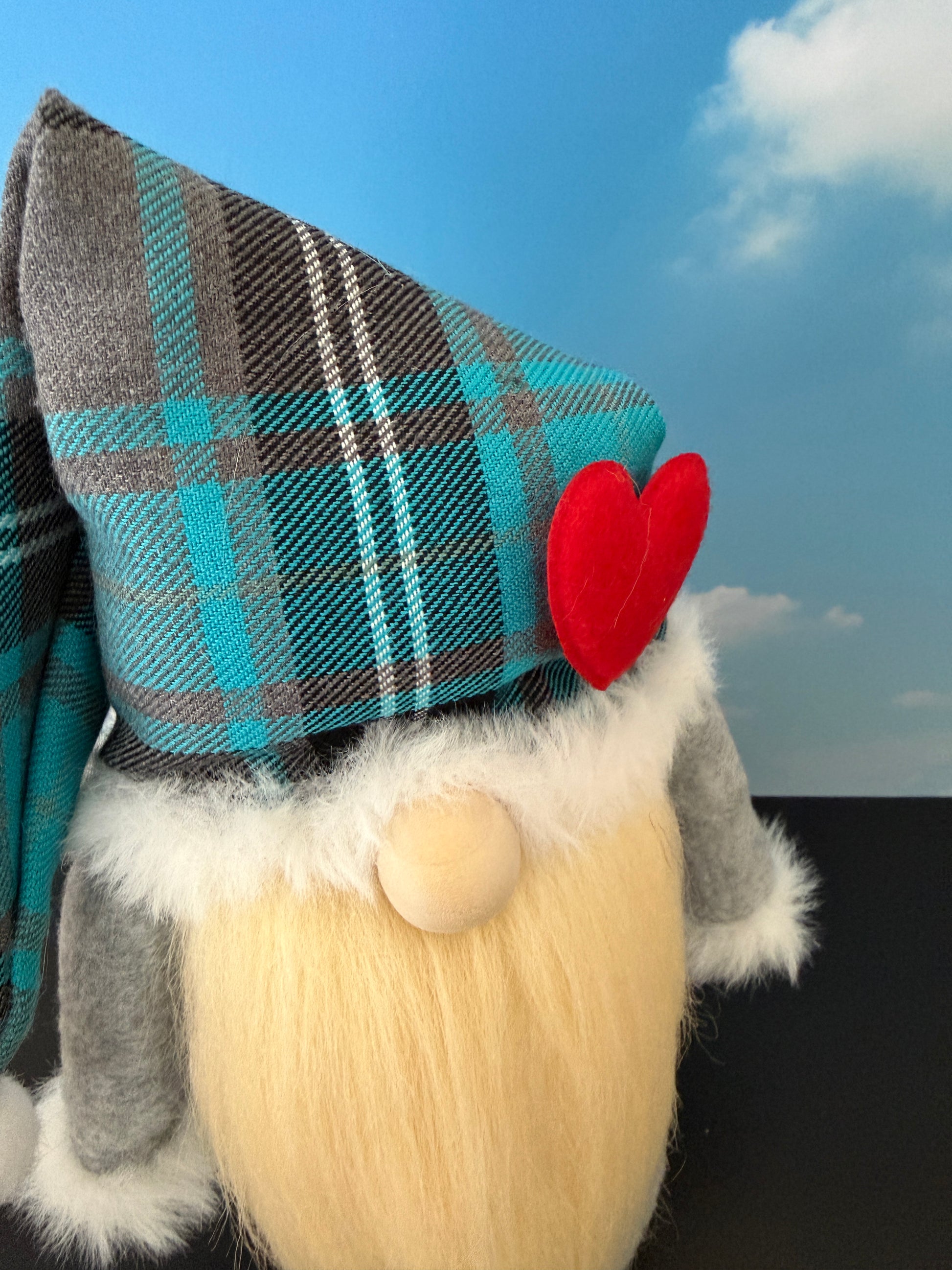 A close-up of a Northern Nomes handmade gnome wearing a teal, gray, and white plaid pointed nightcap with a red heart accent, a cream beard, and gray sleeves, set against a blue sky backdrop.