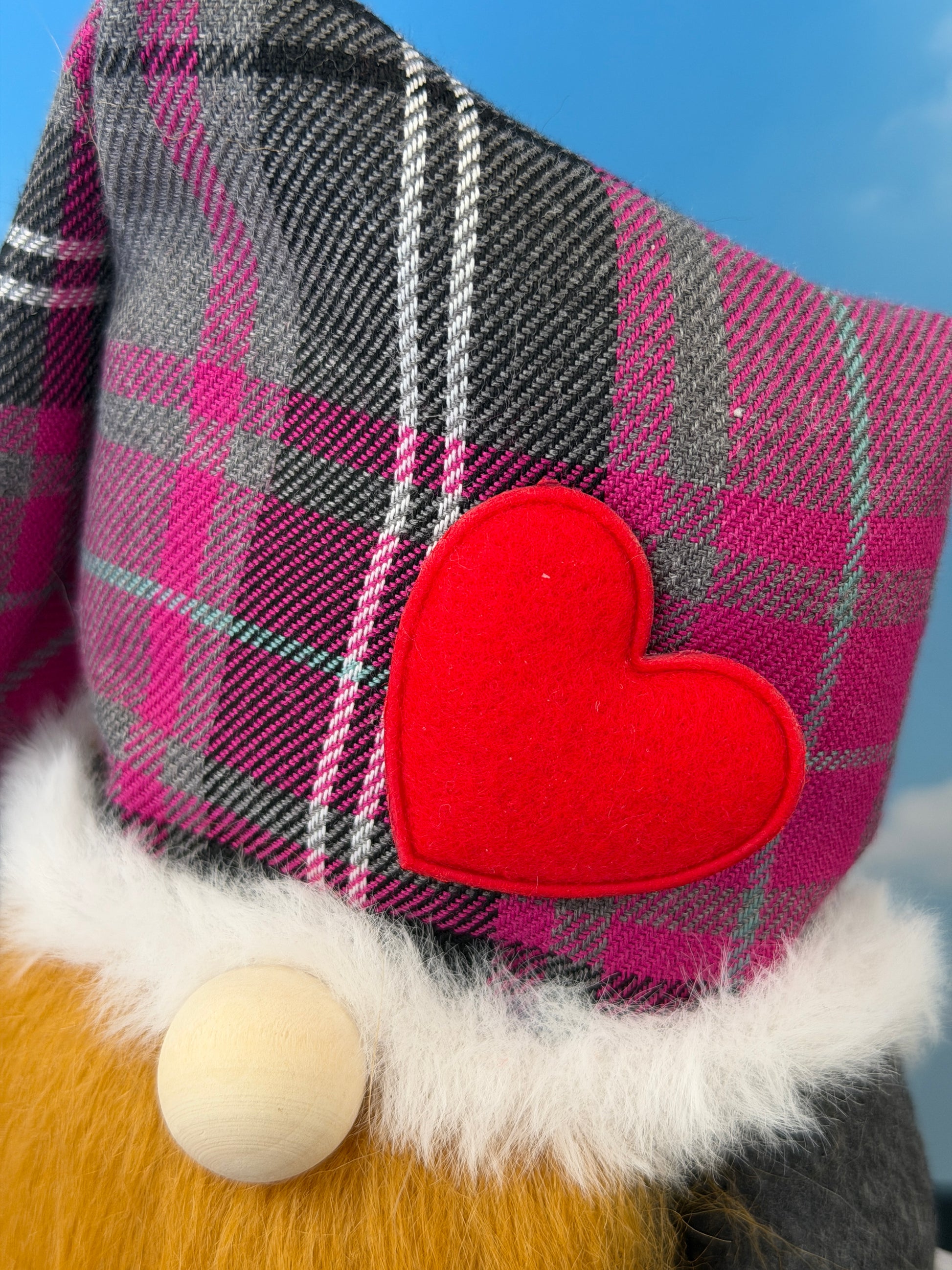 A close-up of a gnome hat in a purple, grey and white plaid fabric featuring a bright red felt heart applique and white faux fur trim, handmade by Northern Nomes.
