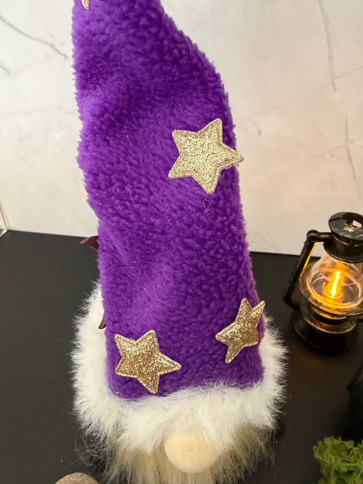 A purple hat on a wizard gnome with three glittery gold stars and a white faux-fur trim, shown close up on a dark tabletop with a lantern in the background.