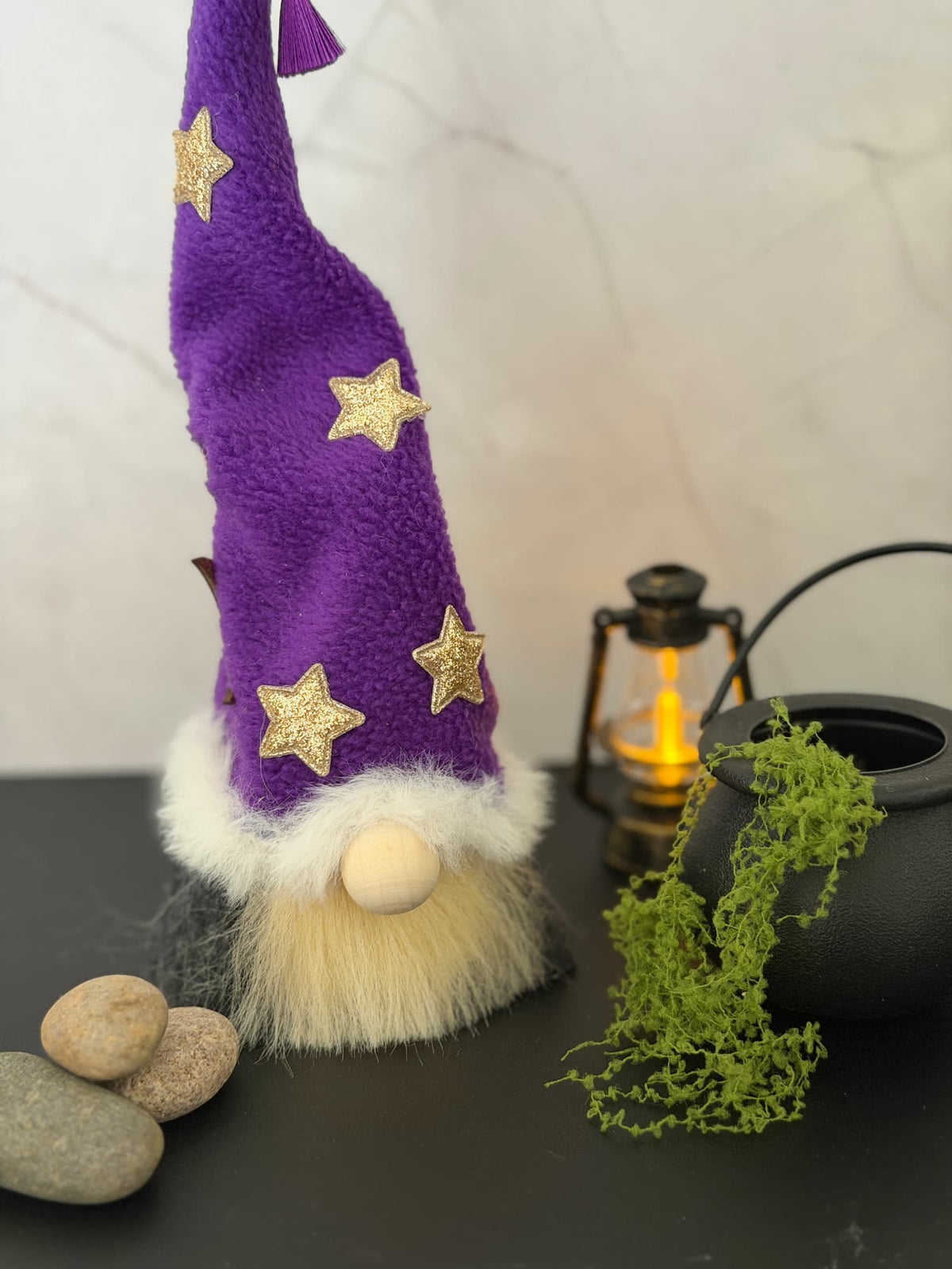 A decorative gnome wizard figurine with a tall purple hat adorned with gold stars, a fluffy cream beard, and a dark body, styled on a tabletop with stones, moss, and a lantern against a light marble wall.
Handmade by Northern Nomes.
