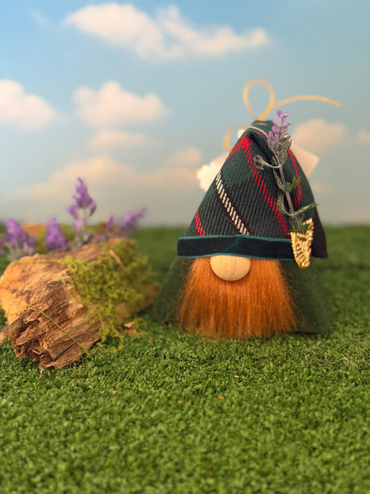 A small gonk with a fluffy ginger beard and a dark tartan hat decorated with a purple heather flower and gold charm, standing on artificial green grass with a log and purple flowers against a blue sky backdrop. Handmade by Northern Nomes.