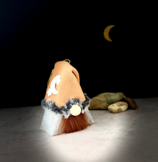 A small gnome figurine with a tan hat, white beard, dark grey faux-fur trim, and dark brown beard, shown on a light surface against a black background with a crescent moon and small stones. Part of the Monnshadow collection, handmade by Northern Nomes.