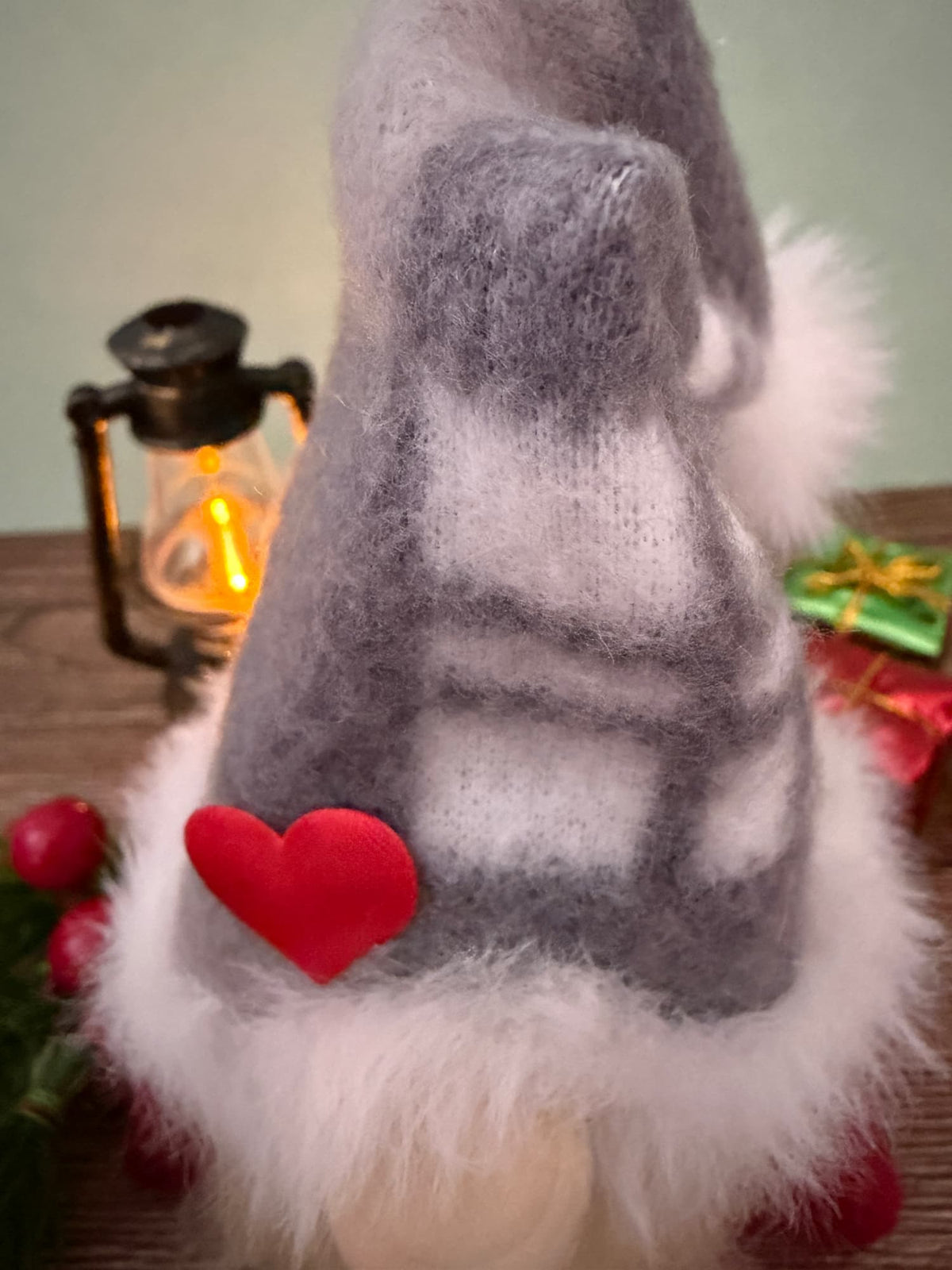 A close-up of a gray fleece gnome hat with white faux fur trim and a red felt heart accent, shown against a softly blurred holiday background with a lantern and wrapped gift.