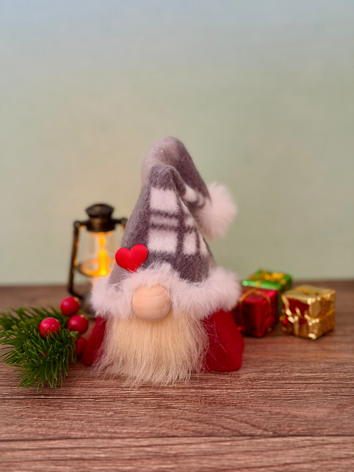 A small Christmas gnome figurine with a gray and white plaid hat, fluffy cream beard, red clothing, and a red heart accent, displayed on a wooden surface with a lantern, pine branch, and wrapped gifts in the background. Handmade by Northern Nomes, part of the Yuletide Folk collection.