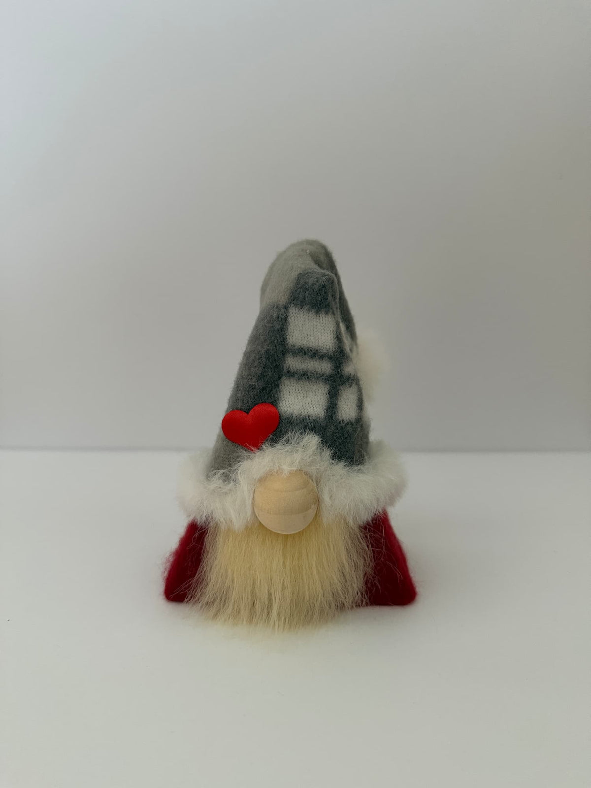 A small handmade gnome figurine with a gray and white plaid hat, a red heart accent, a white faux-fur collar, a beige wooden nose, and a cream beard, set against a plain white background. Handmade by Northern Nomes.