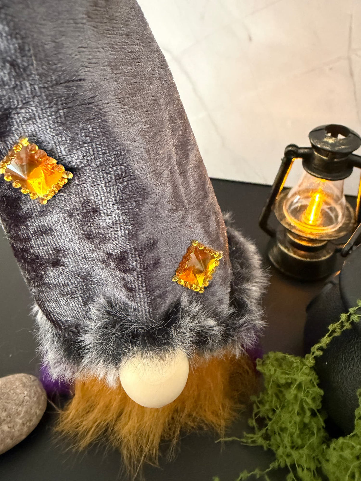 A close-up of a decorative gnome figure with a dark grey velvet hat, two amber gem accents, a white wooden nose and orange-brown faux fur beard, styled on a tabletop with a lit lantern, moss, and stones.