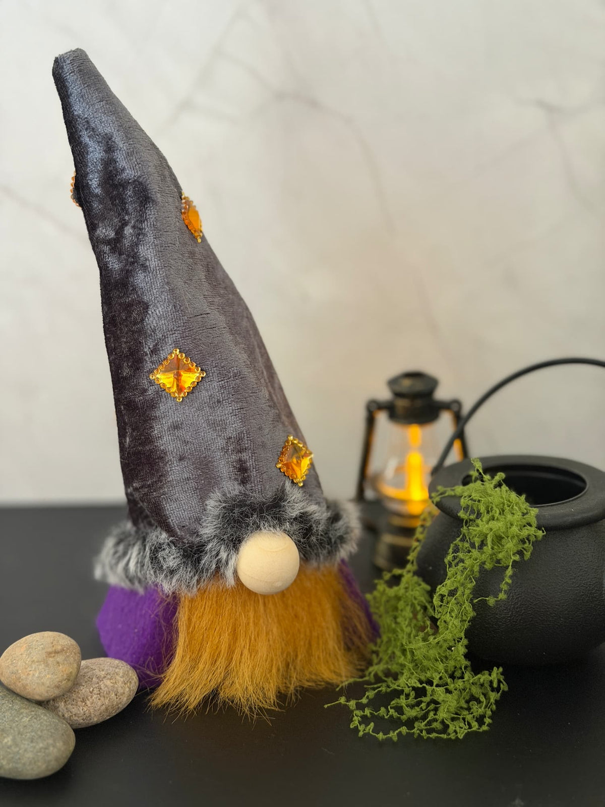 A small decorative gnome wizard figurine with a tall dark gray velvet hat, ginger faux fur beard, purple body and a round white wooden button nose, styled on a dark tabletop with stones, a lantern, greenery, and a black pot against a light marble wall. Handmade by Northern Nomes.
