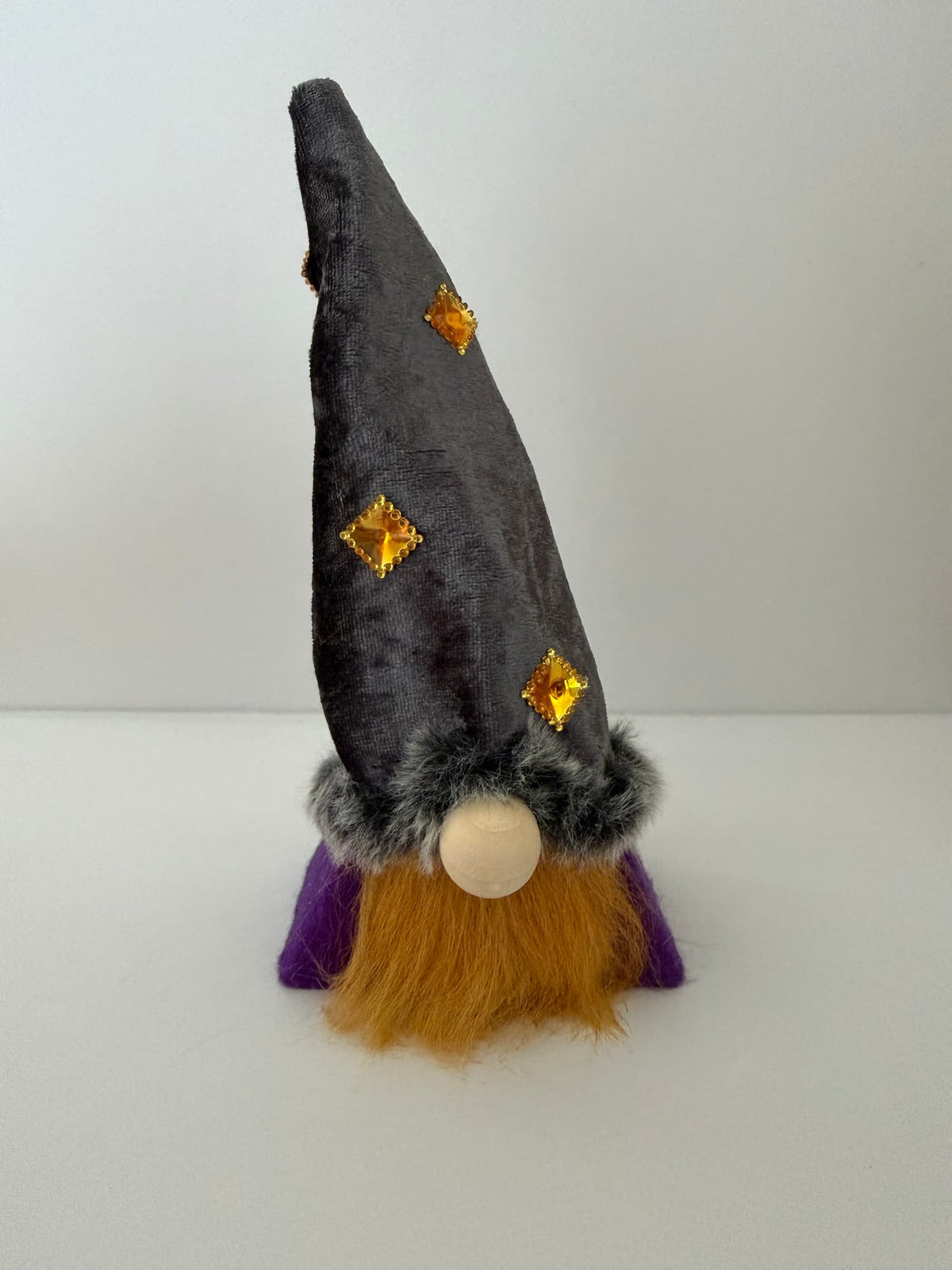A small wizard gnome figurine with a tall dark gray velvet pointed hat decorated with gold diamond-shaped accents, a fluffy orange beard, a purple robe, and a grey faux-fur trim around the hat, shown against a plain light background. Handmade by Northern Nomes.