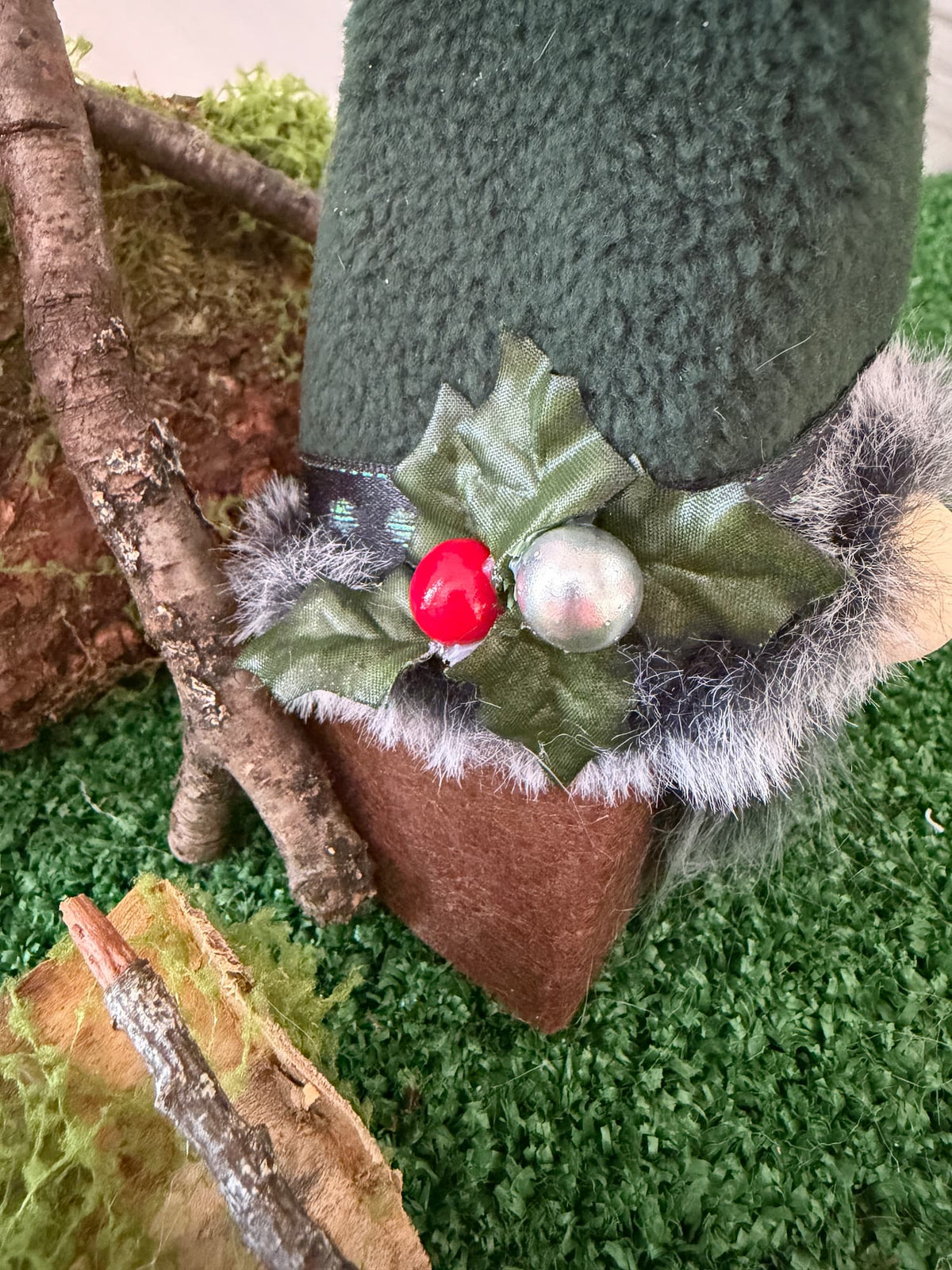 A close-up of a green felt gnome hat decorated with faux holly leaves, a red berry, a silver bead, and gray faux fur trim, set against artificial grass and rustic logs.