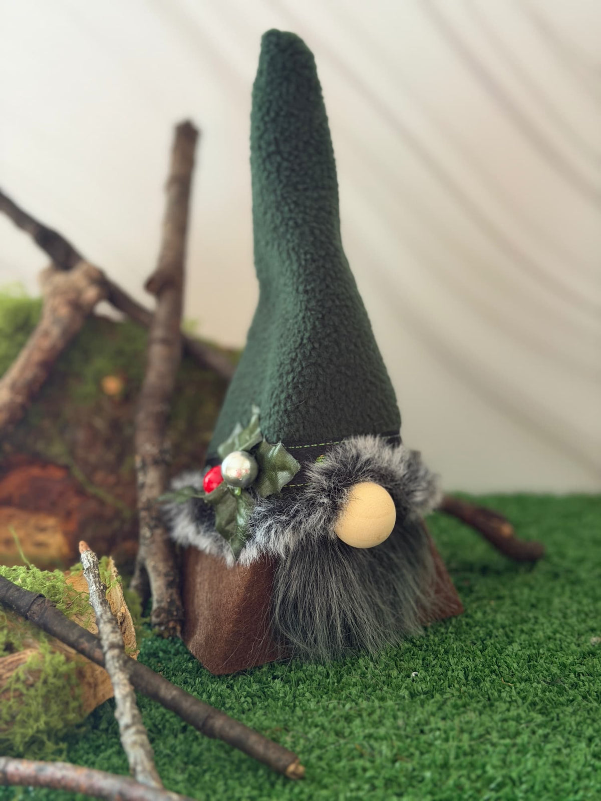 A small gnome decoration with a tall dark green fleece hat, gray faux-fur beard, wooden base, and a beige round nose, styled on green artificial turf with branches and moss. Handmade by Northern Nomes.