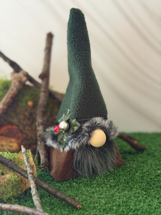 A small gnome decoration with a tall dark green fleece hat, gray faux-fur beard, wooden base, and a beige round nose, styled on green artificial turf with branches and moss. Handmade by Northern Nomes.