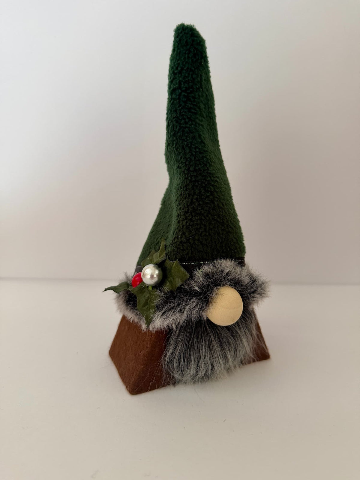 A small gnome decoration with a tall dark green felt hat, gray faux fur beard, wooden nose, and brown base, accented with holly and a pearl bead.