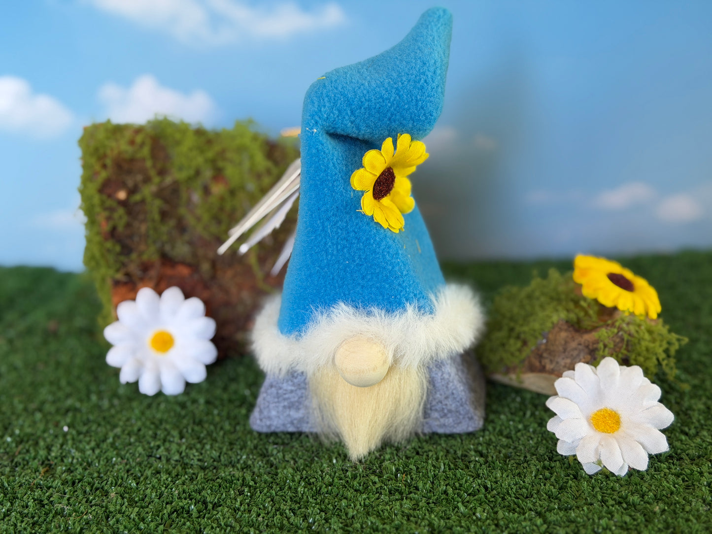 A small felt gnome decoration with a bright blue pointed hat, fluffy cream beard, and a yellow sunflower accent, displayed on artificial grass with daisies and mossy rocks against a blue sky backdrop. Bloom Blossom handmade spring folk ornament