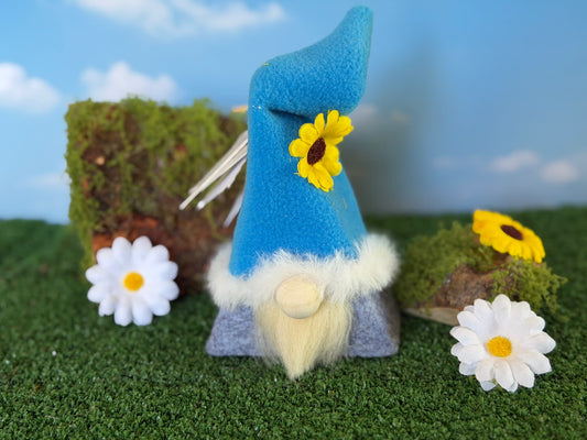 A small handmade gnome decoration with a bright blue pointed hat, fluffy cream beard, and gray body, styled on artificial grass with white daisies, mossy rocks, and a blue sky backdrop. From the Blossom Folk range by Northern Nomes.