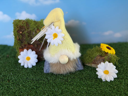 A small decorative gnome figurine with a fleece yellow hat, white daisy flower, and a gray beard, displayed on artificial grass with mossy props and a blue sky backdrop. Handmade by Northern Nomes.