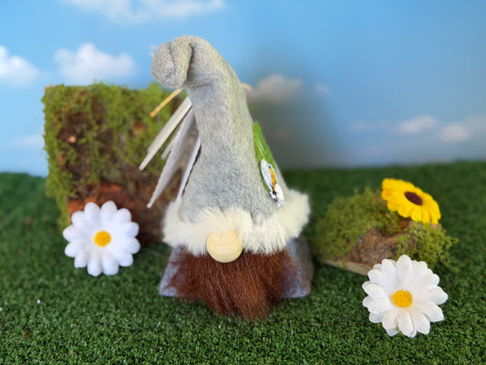A small handmade gnome character with a gray hat, white faux fur trim, brown beard,, posed on artificial grass with daisies, a mossy log, and a blue sky backdrop. Made by Northern Nomes.