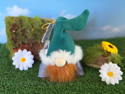 A small plush gnome figurine with a teal hat, orange beard, white fluffy trim, and gray body, styled on artificial grass with white daisies, mossy rocks, and a blue sky backdrop. Handmade character gonk by Northern Nomes.