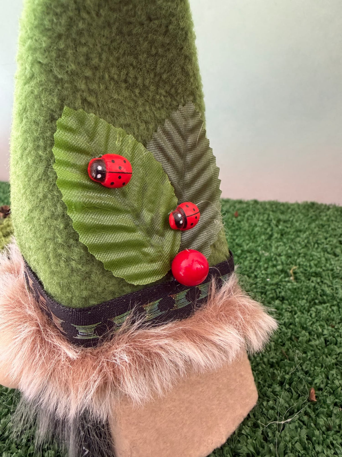A close-up of a green fleece gnome hat decorated with two green fabric leaves, twored ladybug embellishments, and a faux fur trim at the brim, shown standing on artificial grass.