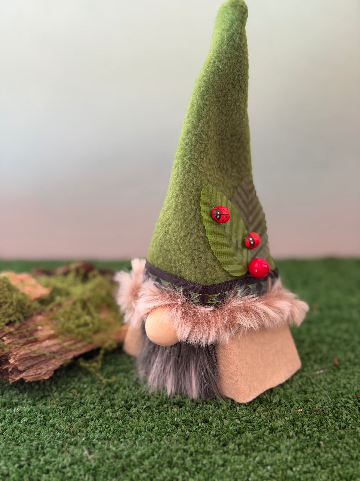 A small decorative gnome figurine with a tall green fleece hat with a beige faux fur trim and grey beard, standing on artificial green grass with a mossy log prop against a soft neutral background. Handmade by Northern Nomes.