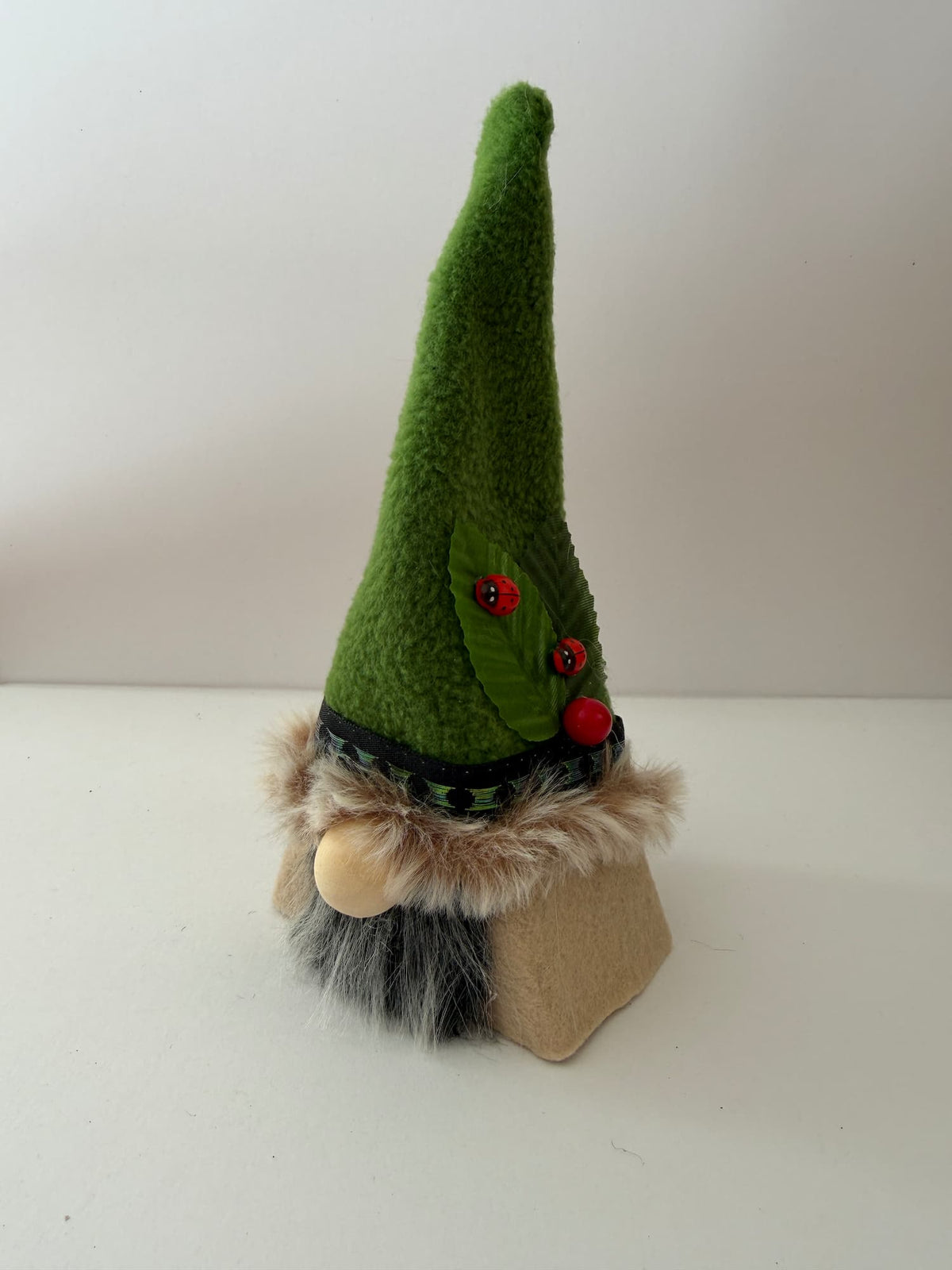 A handmade gnome figurine with a tall green pointed hat decorated with a green leaf and red berry and two ladybirds, a beige wooden bead nose, a grey and black faux fur beard, and a tan base, against a white background.
