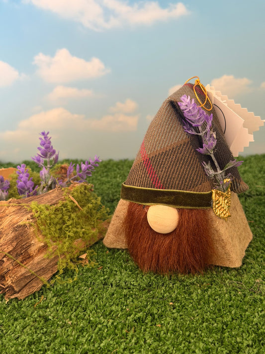 A decorative gnome figurine with a brown plaid hat, brown faux fur beard, and purple lavender accents, displayed on artificial grass next to a mossy log prop under a blue sky backdrop. Handmade with reclaimed timber covered with felt made by Northern Nomes.