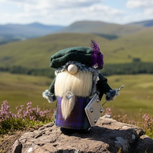 A handmade Scottish-style gnome figurine wearing a dark green kilt, purple feathered bonnet, and long blond beard, standing on a rock with a blurred mountain landscape in the background. Carrying a sword and a shield. Made by Northern Nomes.