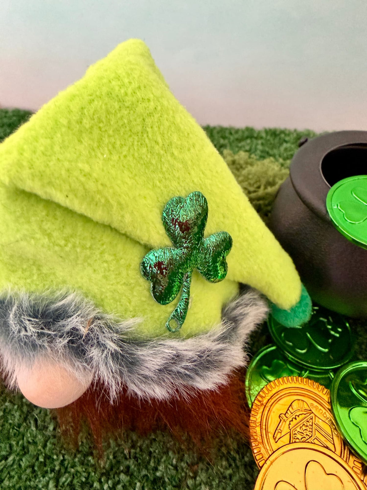 A close-up of a St. Patrick's Day gnome decoration wearing a bright lime green felt hat with a glittery green shamrock accent, gray faux fur trim, and a brown beard, styled with a black cauldron and gold and green coins on artificial grass.