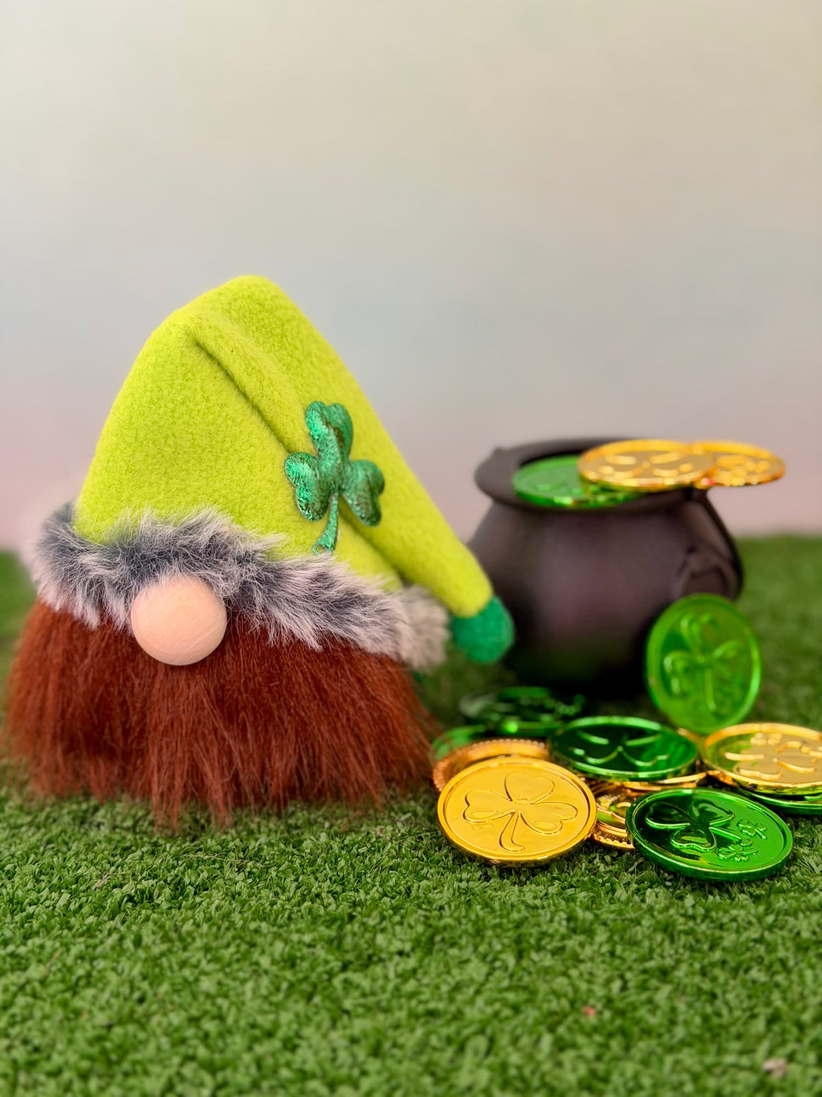 A St. Patrick's Day decorative lucky leprechaun handmade gnome figurine with a bright green felt hat, brown faux-fur beard, gray faux-fur trim, and a wooden button nose, posed beside a black pot and shiny gold and green coins on artificial grass. Made by Northern Nomes.