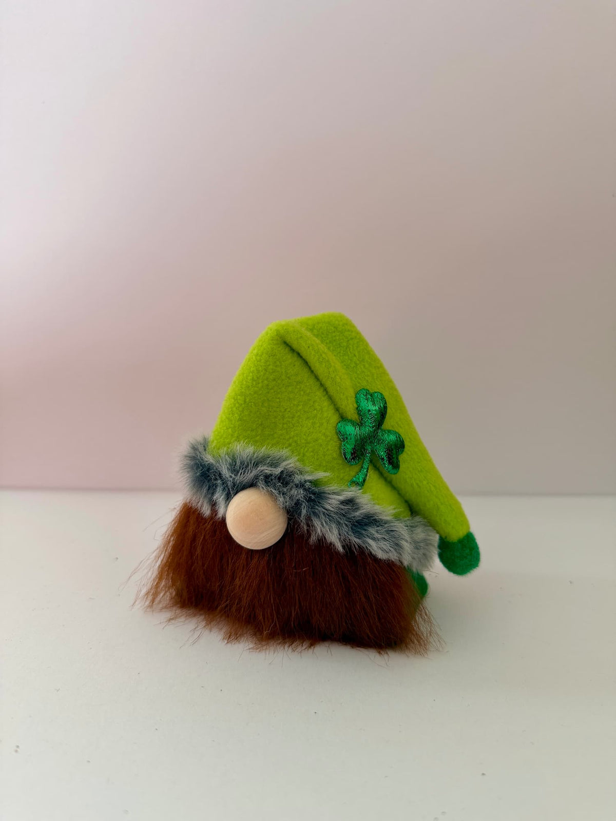 A handmade small lucky leprechaun gnome wearing a bright green pointed hat with a green shamrock, brown faux-fur beard, and a green faux-fur trim, photographed against a plain light background. Part of the Cloverhill Folk collection by Northern Nomes.