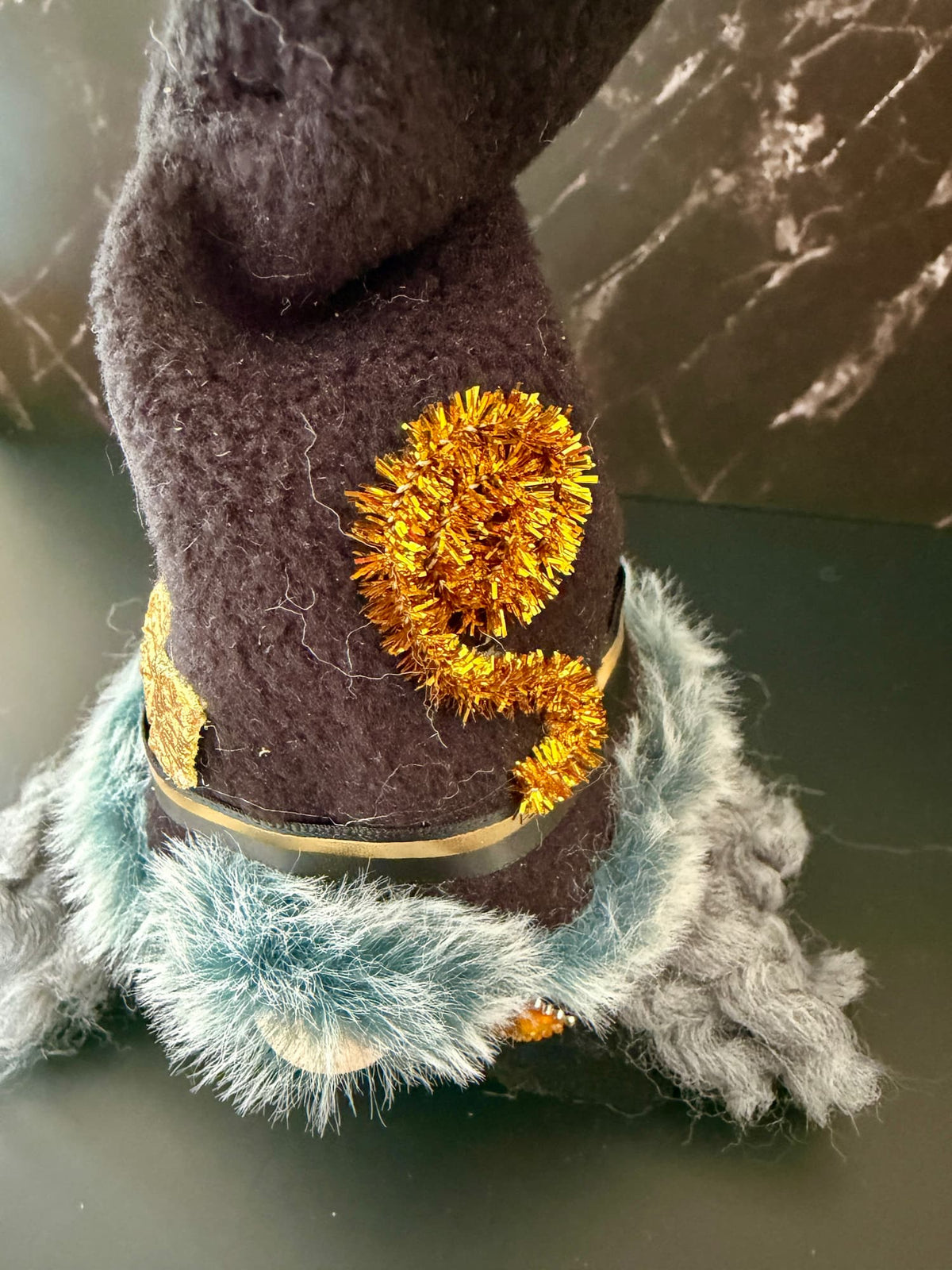 A close-up of a dark brown felted wool hat with a fluffy light blue and gray brim, decorated with a gold tinsel pom-pom and a gold band.