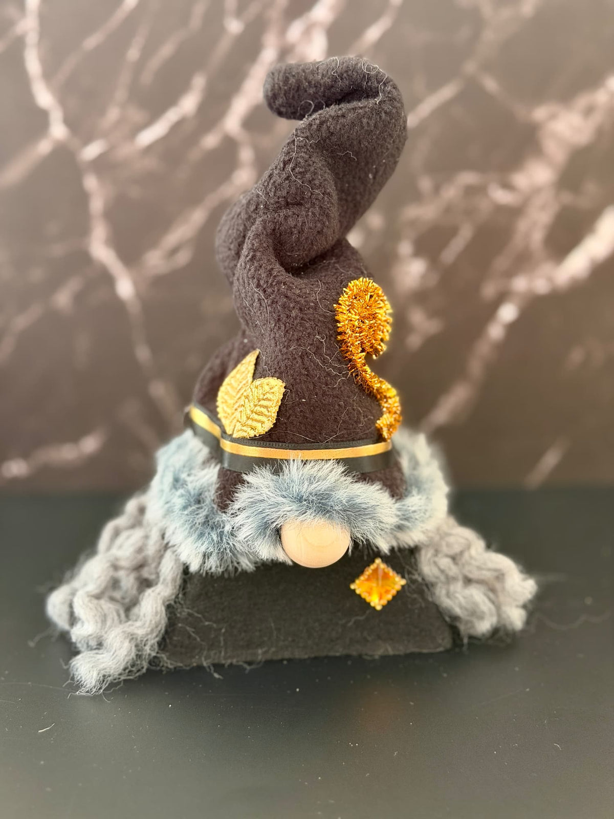A small witch gnome figurine wearing a tall black pointed hat with gold leaf and gold spiral accent, a black body, dark grey curly hair, and a fluffy green grey faux fur trim around the hat, set against a black and marble effect background. Handmade by Northern Nomes.