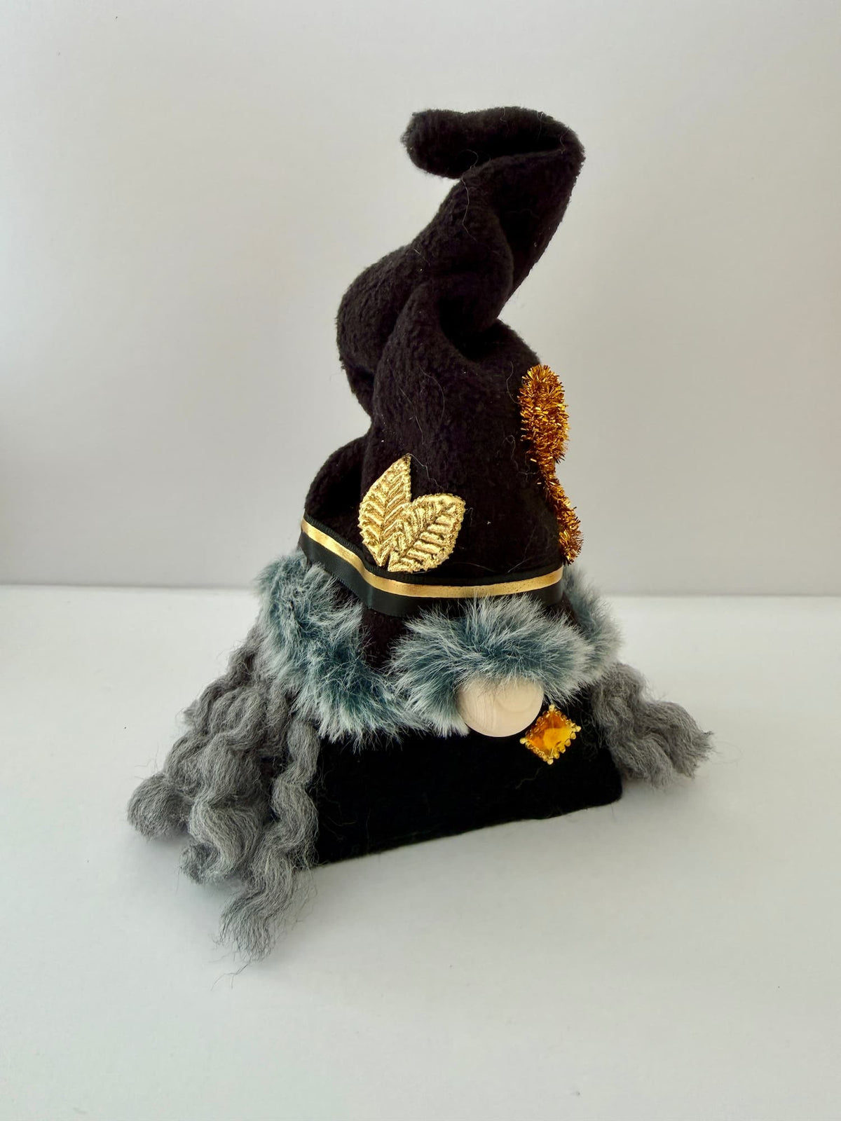 A small witch gnome figurine wearing a tall black pointed hat with gold leaf and gold spiral accent, a black body, dark grey curly hair, and a fluffy green grey faux fur trim around the hat, set against a plain white background. Handmade by Northern Nomes.