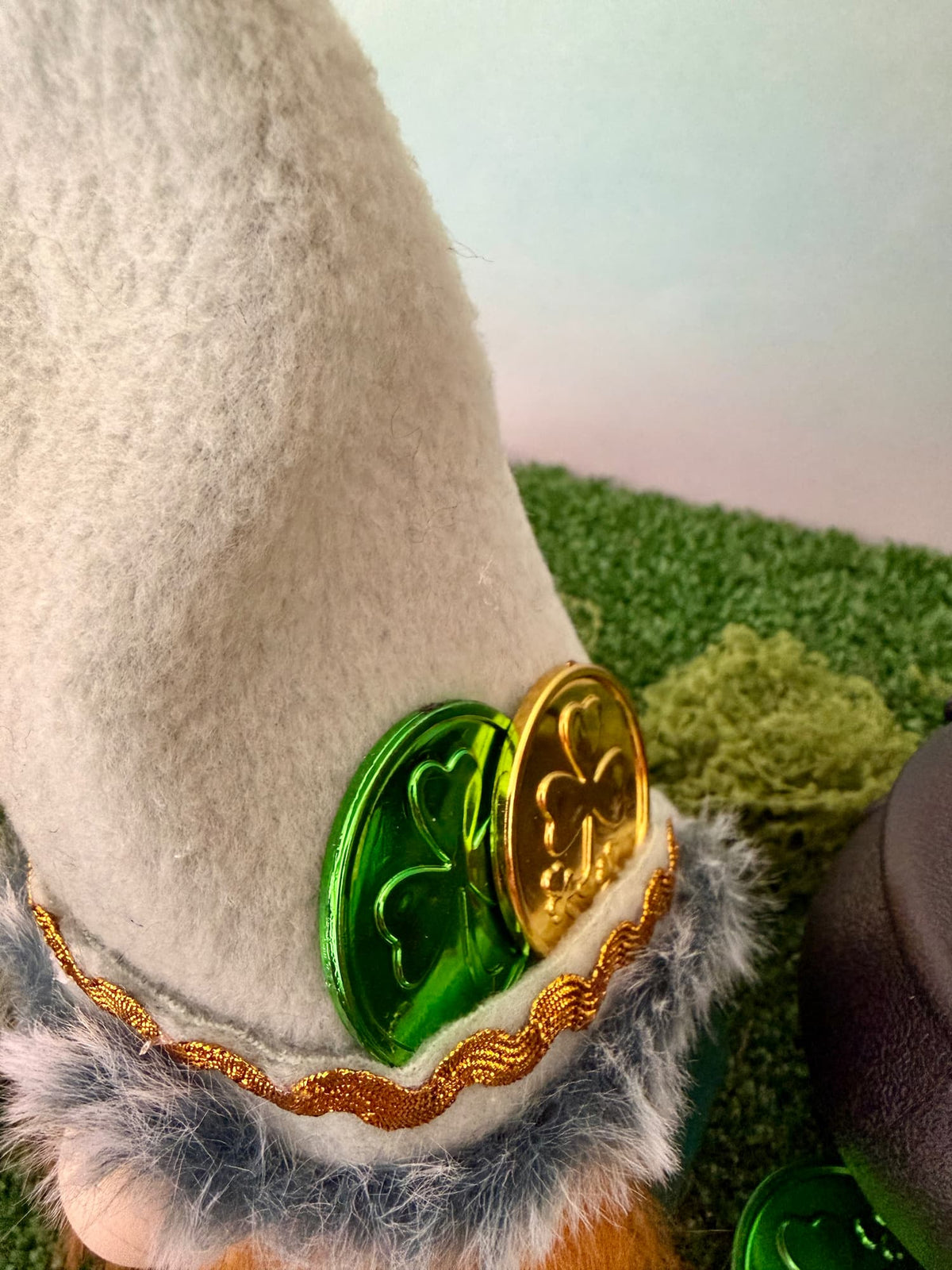 A close-up of a handmade St. Patrick's Day themed decorative gnome hat o featuring a pale green felt hat with faux fur trim, gold and green shamrock coins ornament, and a gold decorative trim.