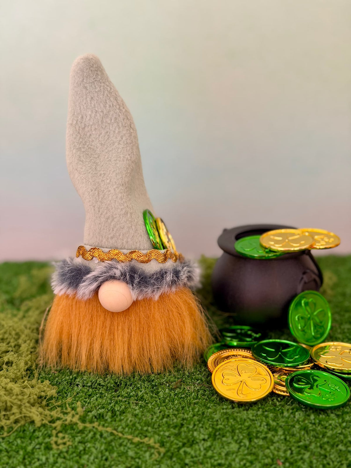 A handmade lucky leprechaun gnome decoration with a tall pale green felt hat, orange faux-fur beard, and greenish faux-fur trim, displayed on green artificial grass next to a black pot and shiny gold and green coins. Made by Northern Nomes and part of the Cloverhill collection.
