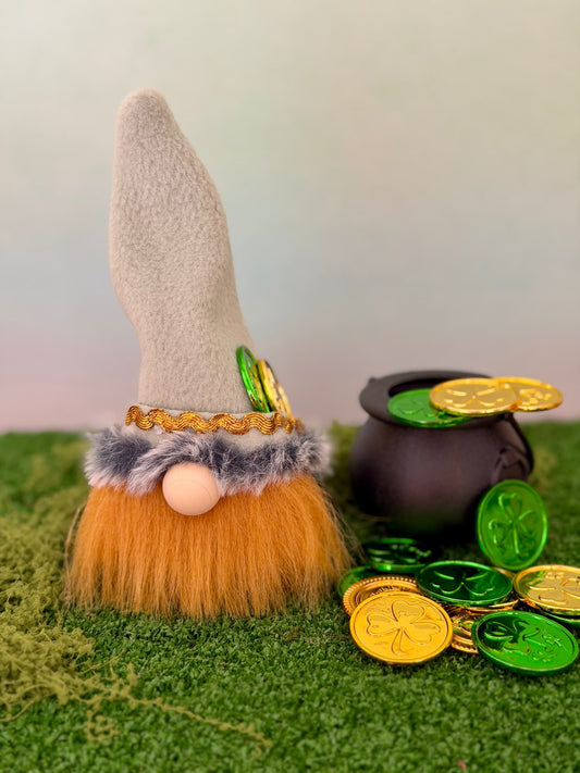 A handmade lucky leprechaun gnome decoration with a tall pale green felt hat, orange faux-fur beard, and greenish faux-fur trim, displayed on green artificial grass next to a black pot and shiny gold and green coins. Made by Northern Nomes and part of the Cloverhill collection.