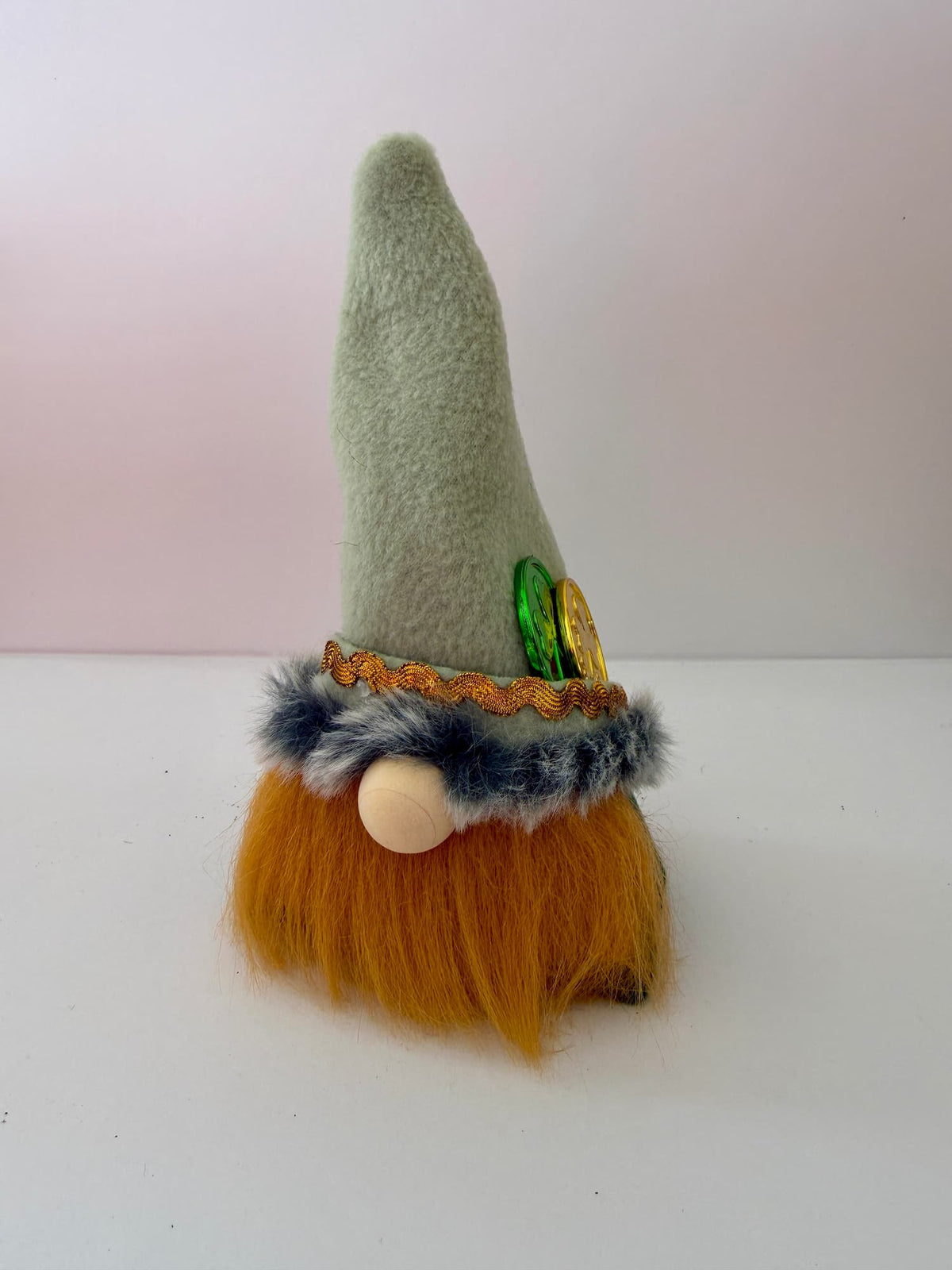 A small handmade St. Patrick's Day gnome figurine with a tall light green felt hat, orange faux-fur beard, blue-gray faux-fur trim, and gold coin accents, photographed on a white background. A lucky leprechaun gnome by Northern Nomes.
