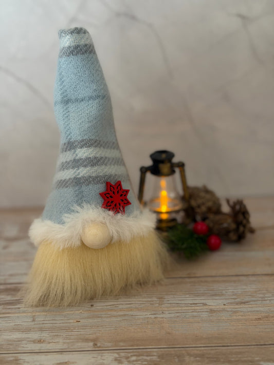 A small Christmas gnome decoration with a light blue and gray striped knit hat, fluffy cream beard, and a red snowflake accent, styled on a rustic wooden surface with a lantern and pinecones in the background. Handmade by Northern Nomes.