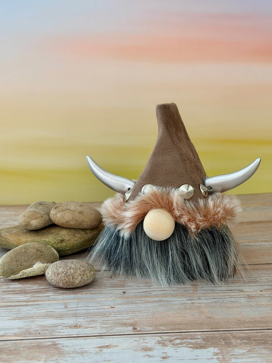 A handmade decorative Viking-style gnome figurine with a brown pointed hat, curved silver horns, a faux-fur hat trim, a round beige wooden nose, and a gray beard, displayed on a wooden surface with stones against a soft pastel background.
