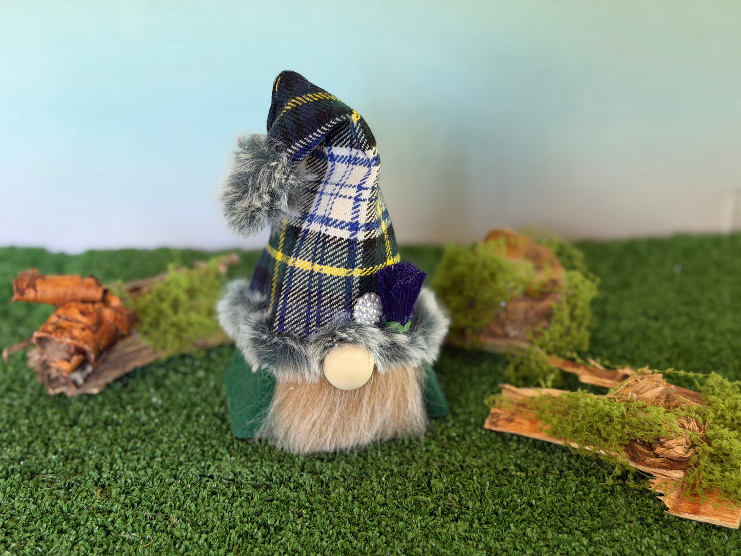 A small decorative gonk figure with a green tartan plaid pointed hat, faux fur trim, a gray beard, and a wooden nose, standing on artificial green turf with moss and wood props against a pale blue background. From the Thistlewood Wee Folk Collection by Northern Nomes 