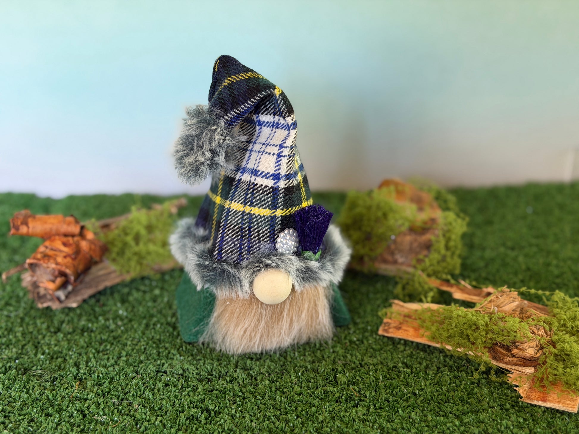 A small decorative gonk figure with a green tartan plaid pointed hat, faux fur trim, a gray beard, and a wooden nose, standing on artificial green turf with moss and wood props against a pale blue background. From the Thistlewood Wee Folk Collection by Northern Nomes 