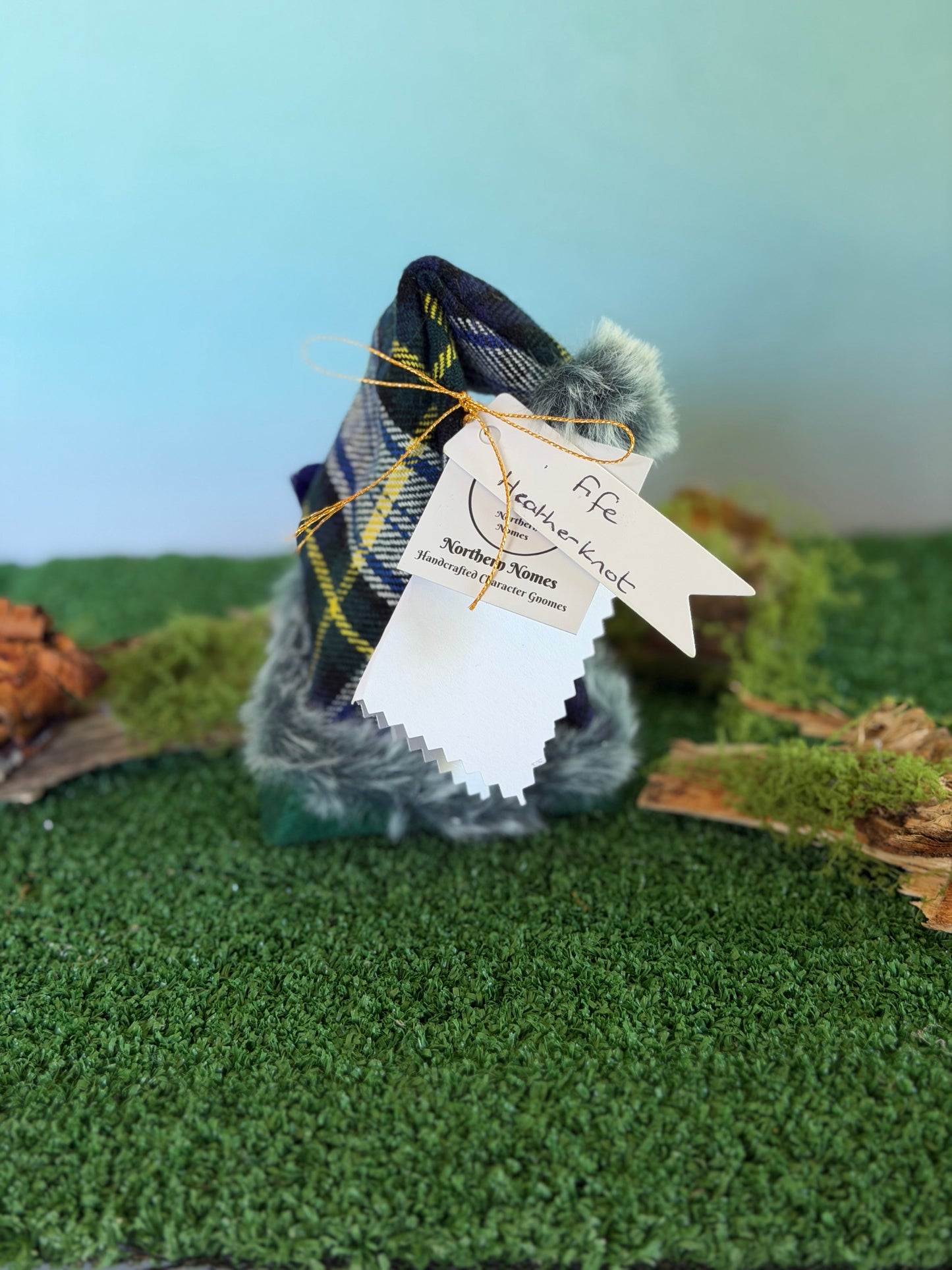 A small handmade gonk figurine dressed in a green tartan plaid top hat with a fluffy pom-pom and a green felt body, displayed on artificial green moss with a light blue background. It has hanging tags, including a tag that reads 'Fife Heatherknot' and a story description card by Northern Nomes.
