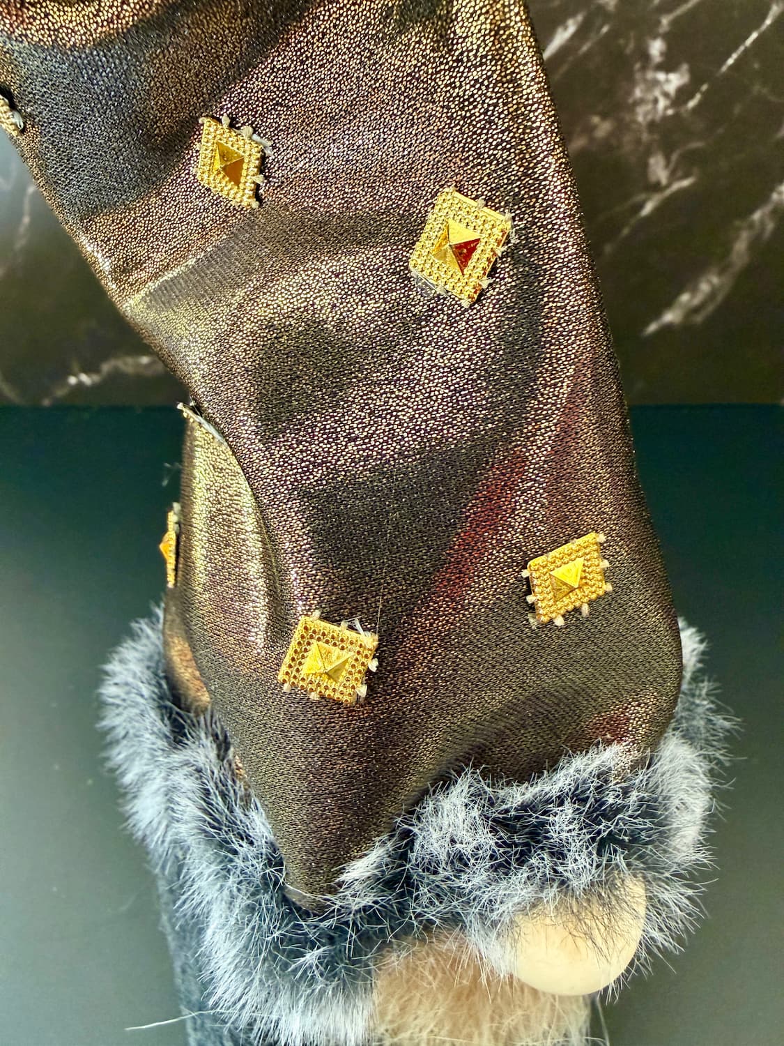 A close-up of a golden textured fabric item with gold diamond-shaped patches and a grey faux-fur trim at the bottom, shown against a dark background.