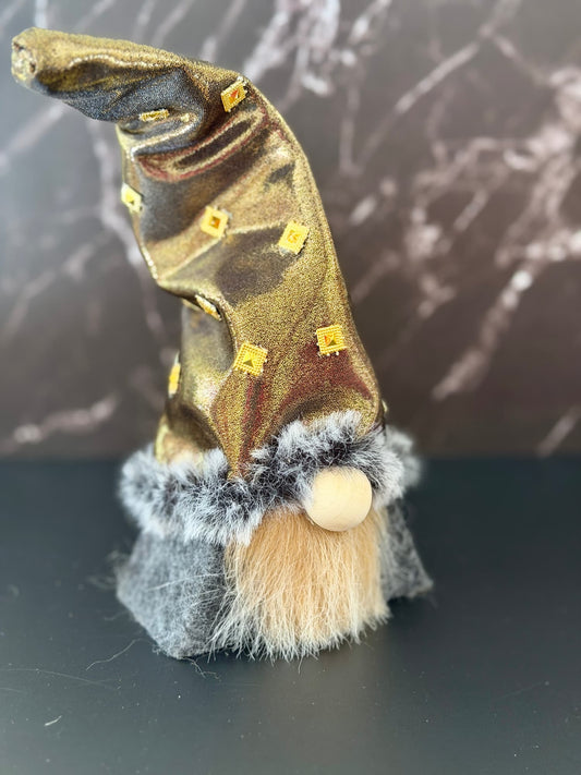 A small gnome figurine with a metallic gold pointed hat decorated with tiny gold star embellishments, a gray faux-fur trim, a beige wooden nose, and a beige beard, standing on a dark surface against a dark marble-patterned background. Handmade by Northern Nomes.