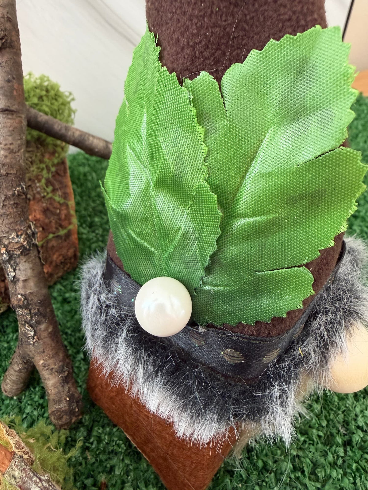 Close-up of a gonk figure wearing a brown hat with two green fabric leaves, a black band, and a white faux-pearl ornament, standing on artificial green turf with a small tree prop.