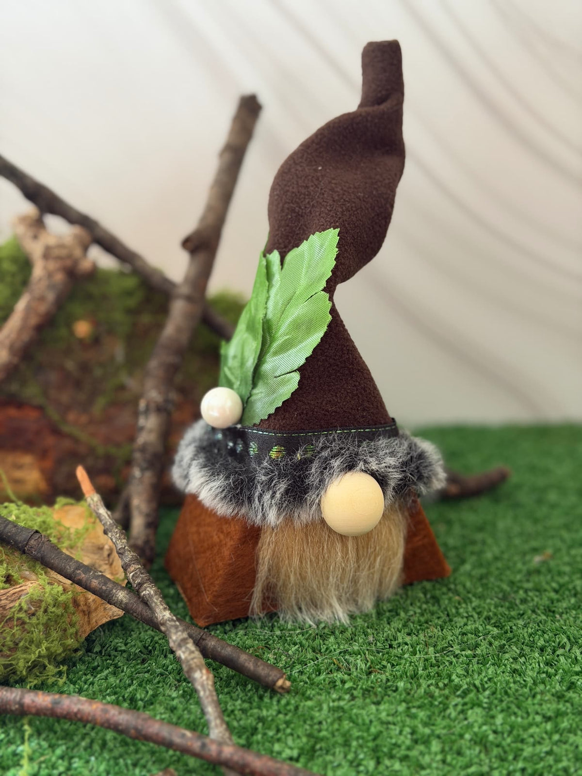 A handmade small forest gnome figurine with a brown fleece hat, green leaf accent, faux fur trim, and a beige beard, displayed on artificial grass with branches and moss.