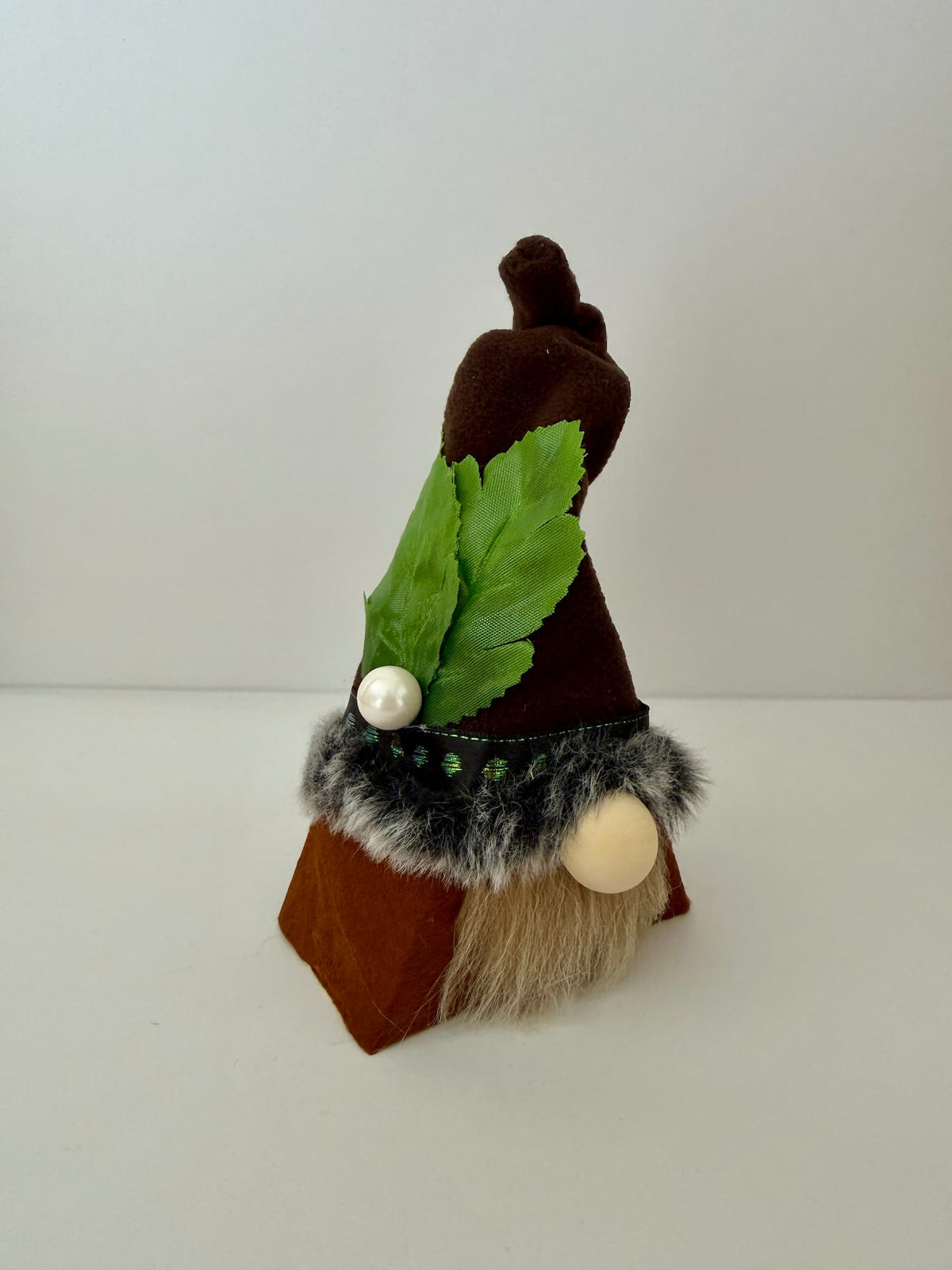 A small decorative gnome figurine with a brown pointed hat, green leaf accent, faux fur trim, and a beige beard, photographed on a plain light background. Handmade by Northern Nomes.